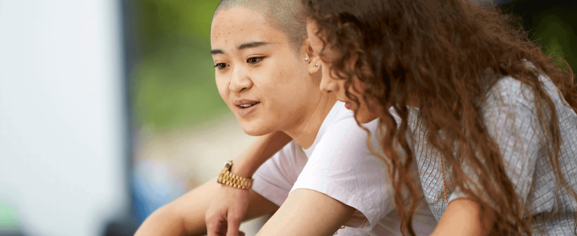 A girl with curly hair has her arm over her girlfriend's shoulder and talks to her while they sit in a park.