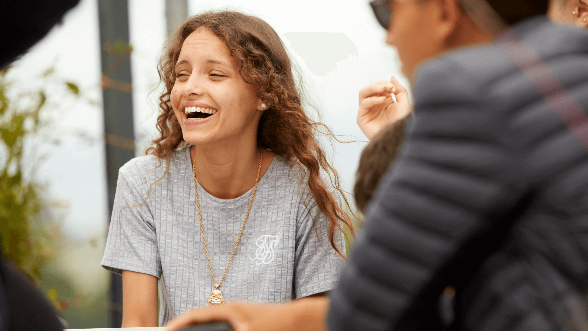 A young woman with long, curly hair wearing a grey T-shirt. She is sitting with her friends and laughing.