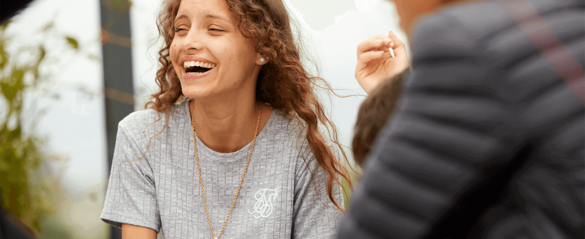 A young woman with long, curly hair wearing a grey T-shirt. She is sitting with her friends and laughing.