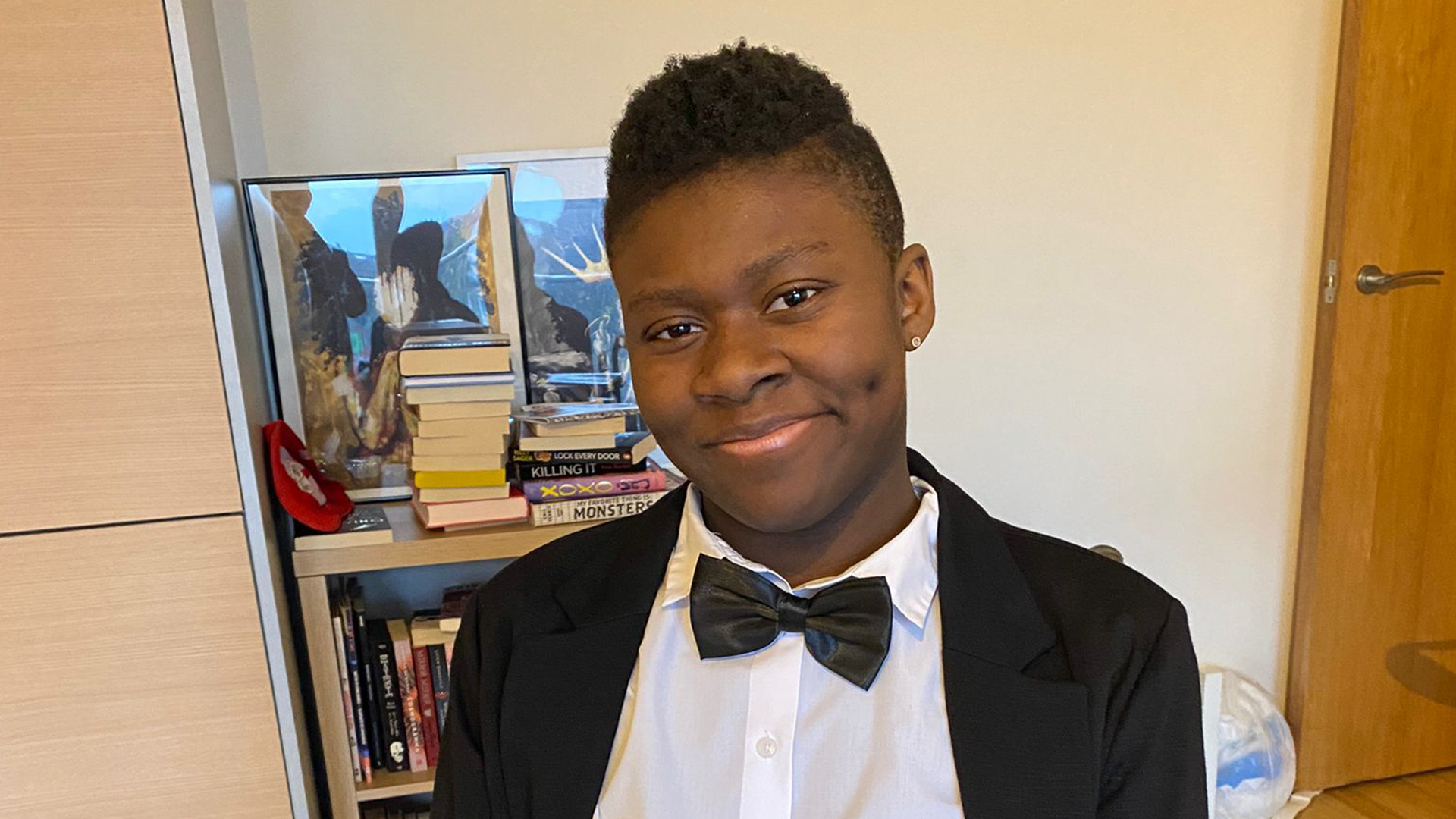Simi is a Black young person and they are wearing a Black tuxedo with bow tie whilst sitting in their wheelchair and smiling.