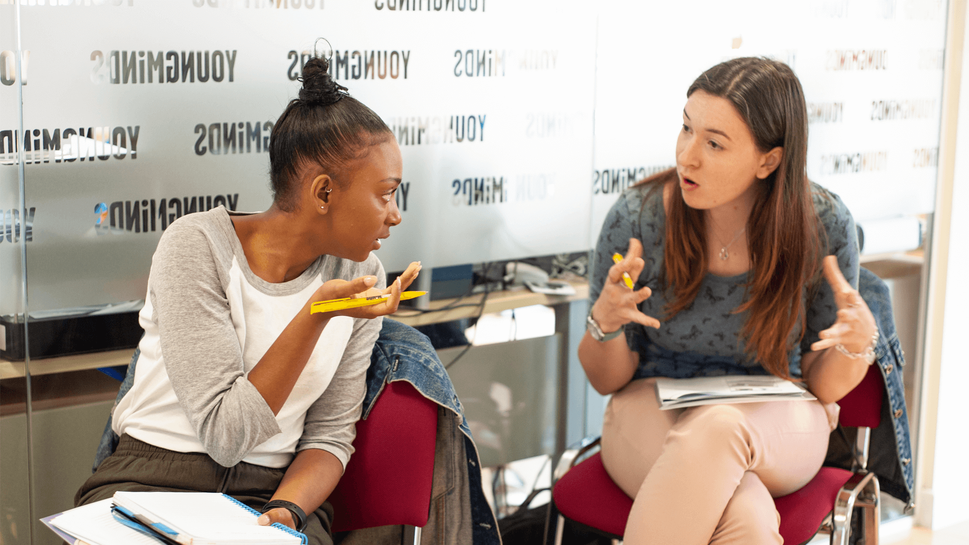 two moms are having a discussion over an activity while sitting on chairs inside a conference room