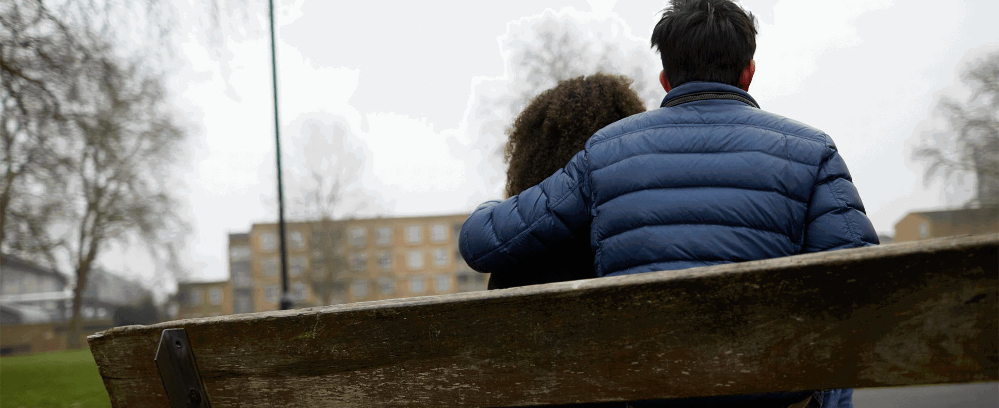 Two young people sitting together on a bench. One has their arm around the other.