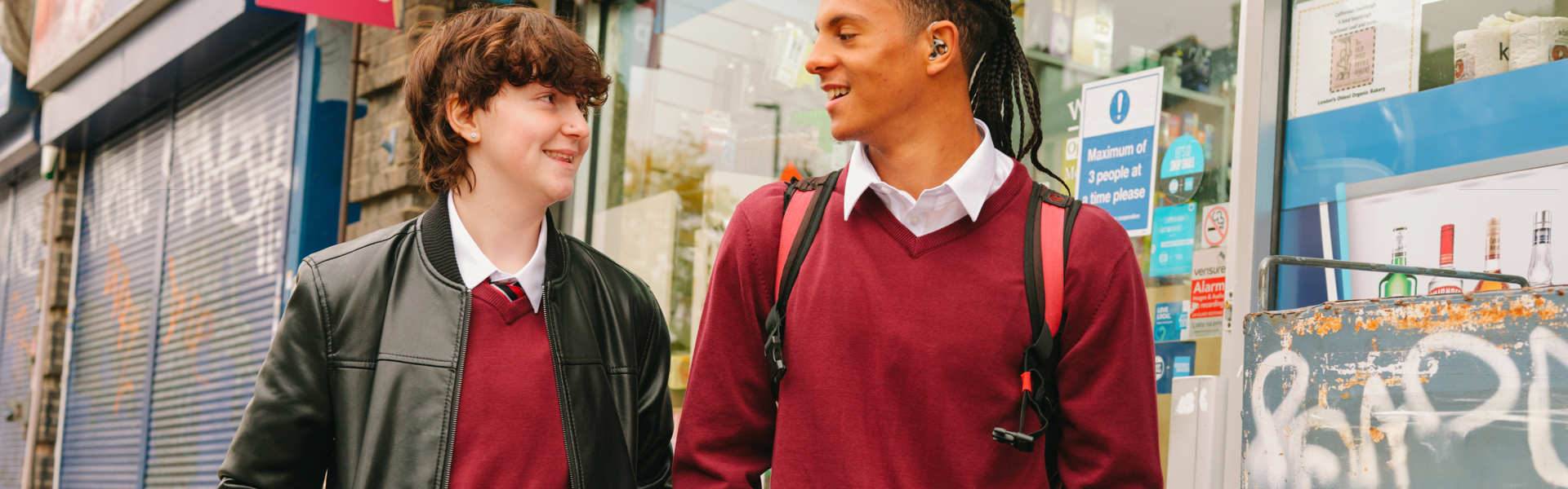 A Black teenage boy wearing a hearing aid speaking to a white non-binary teenager. They are walking on the street outside a shop. Both people are smiling.