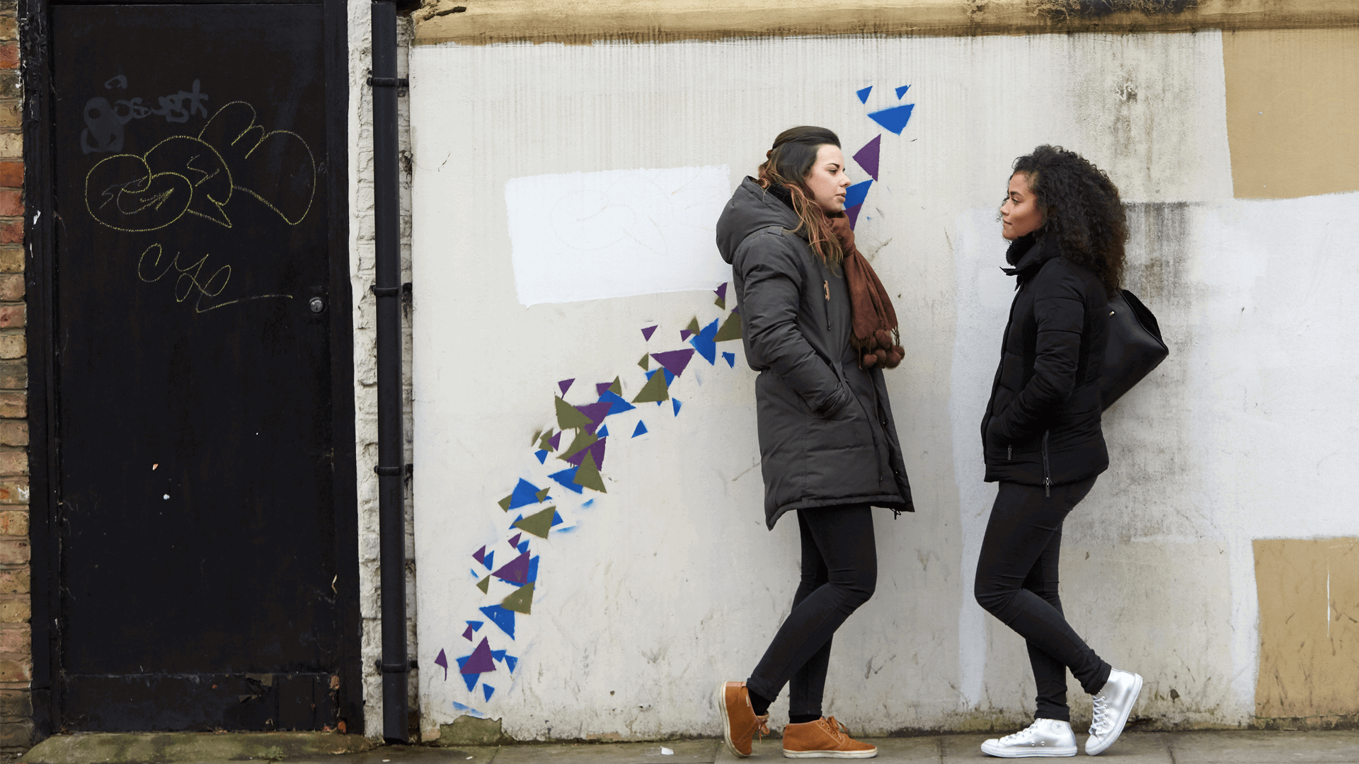 Two girls wearing black jackets are talking while standing against a wall decorated with blue, purple and green graffiti patterns.