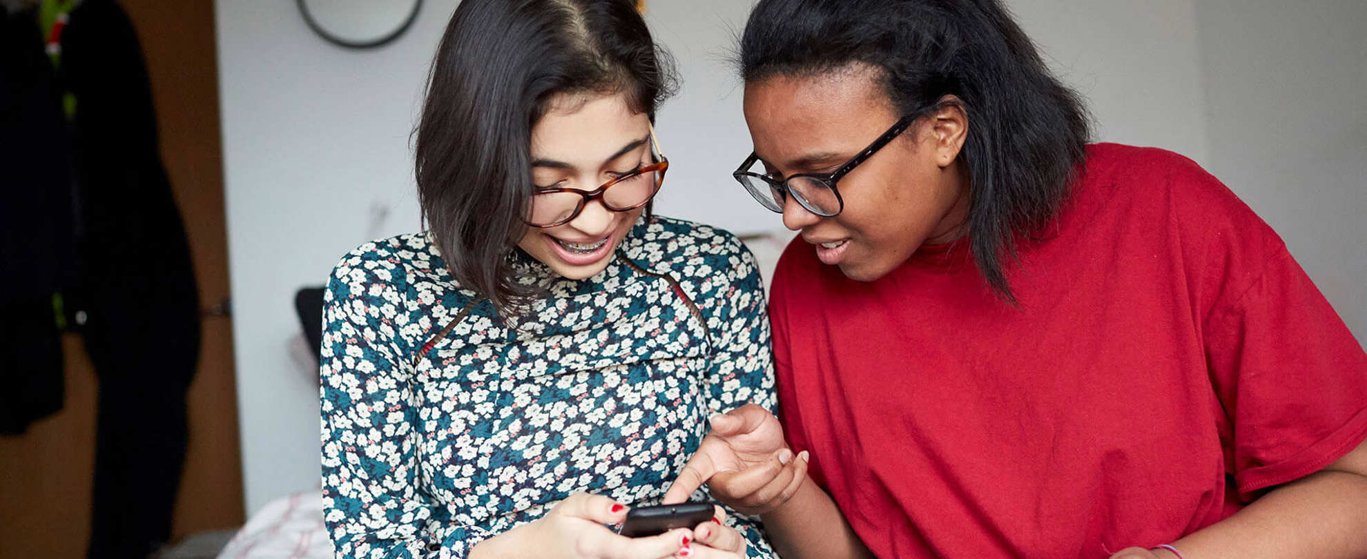 Two young people sitting together on the end of a bed and looking at their mobile phones.
