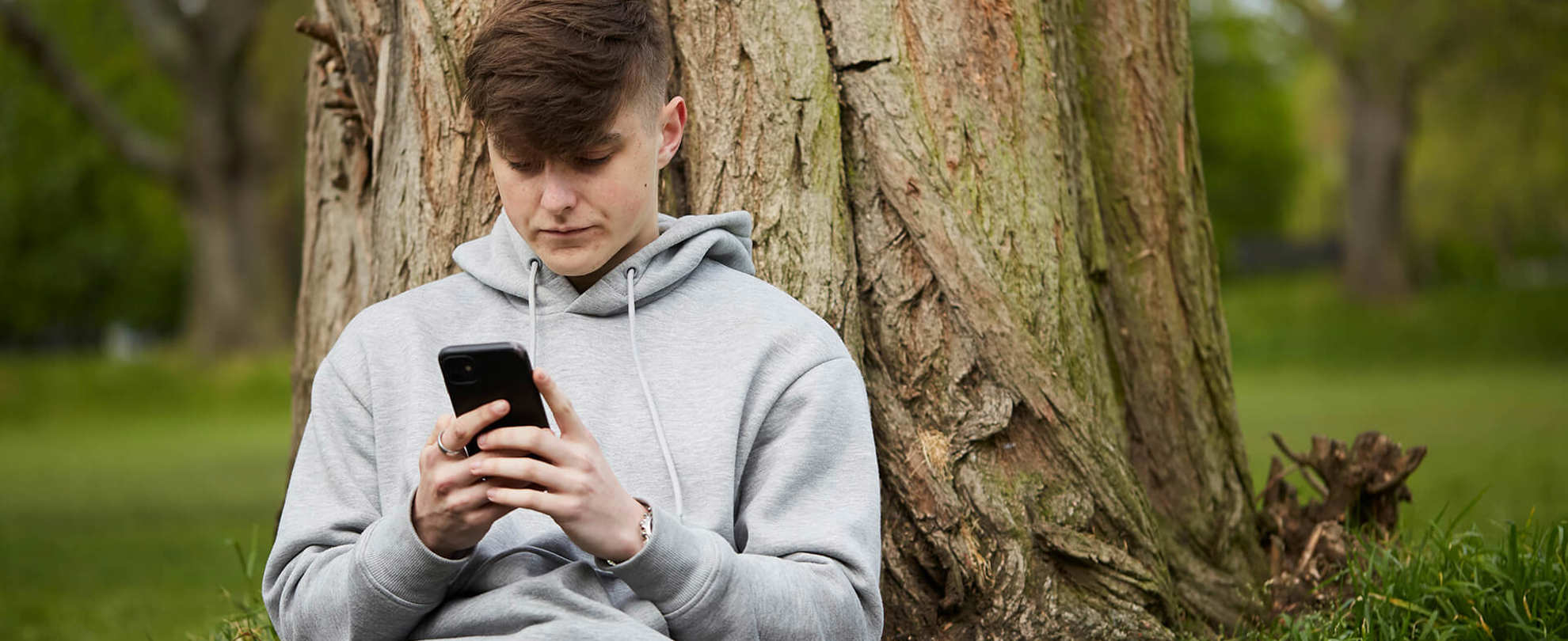 A young man wearing a grey hoodie and looking at his phone while he sits on the grass and leans against a tree.