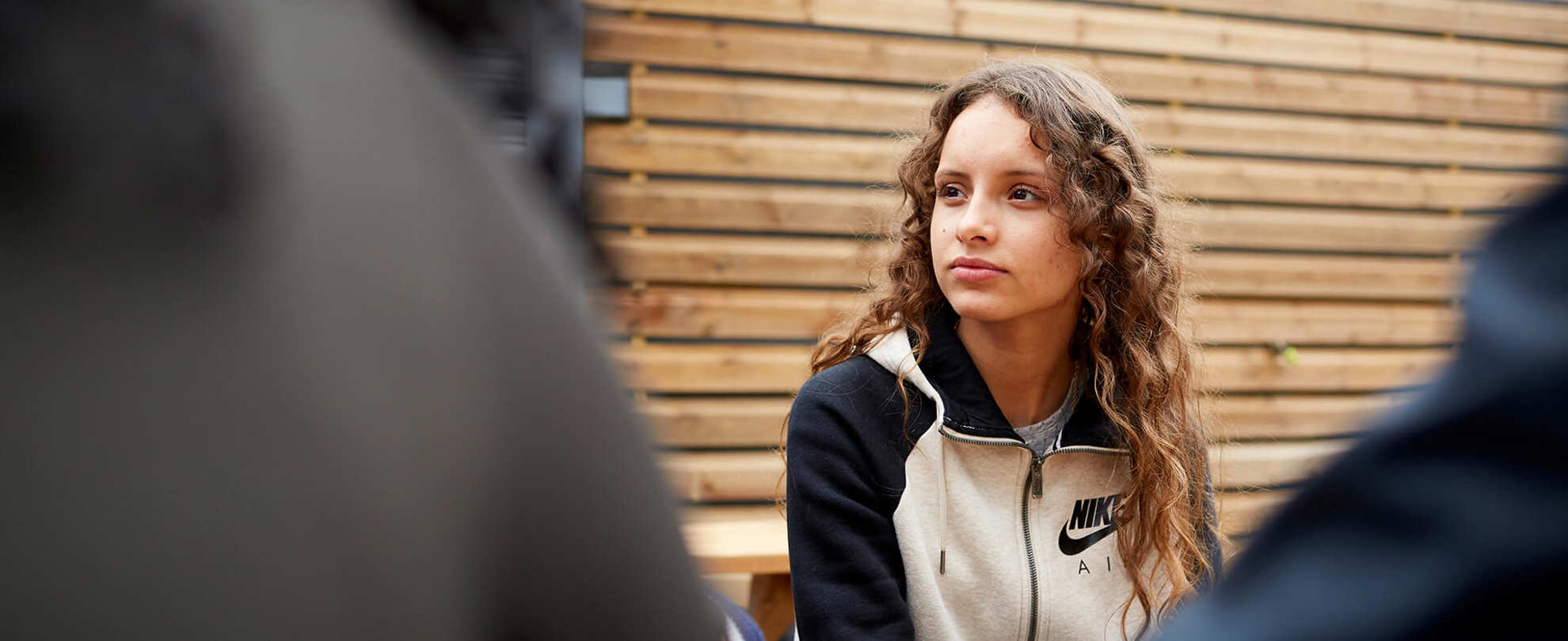 A girl with long, curly hair sits listening to her friends on a picnic bench.
