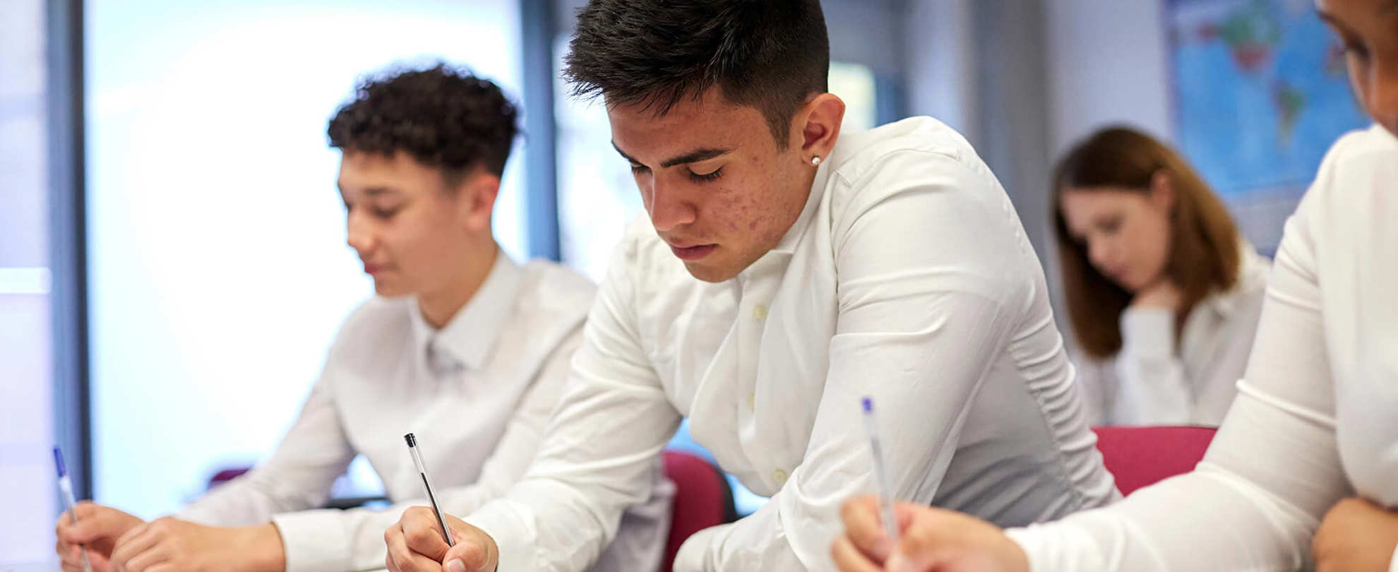 A group of students wearing school uniform sit at their desks in a classroom and write in their textbooks.