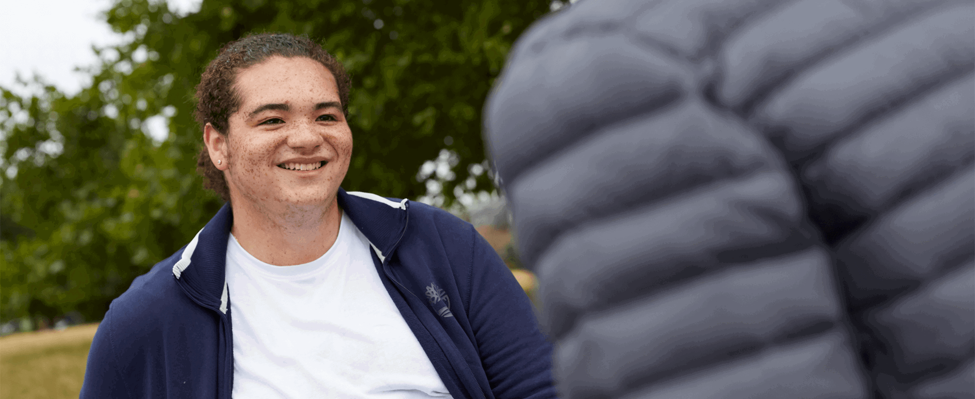 A young person smiling at their friend who is walking towards them.