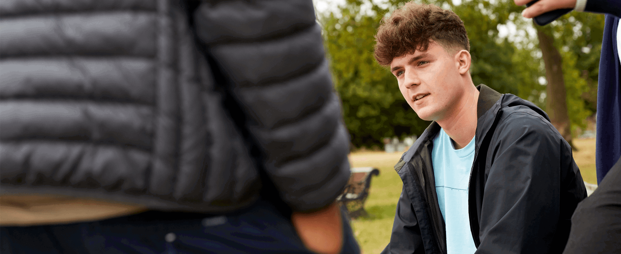 A young person sitting on a bench in a park with friends standing around him.