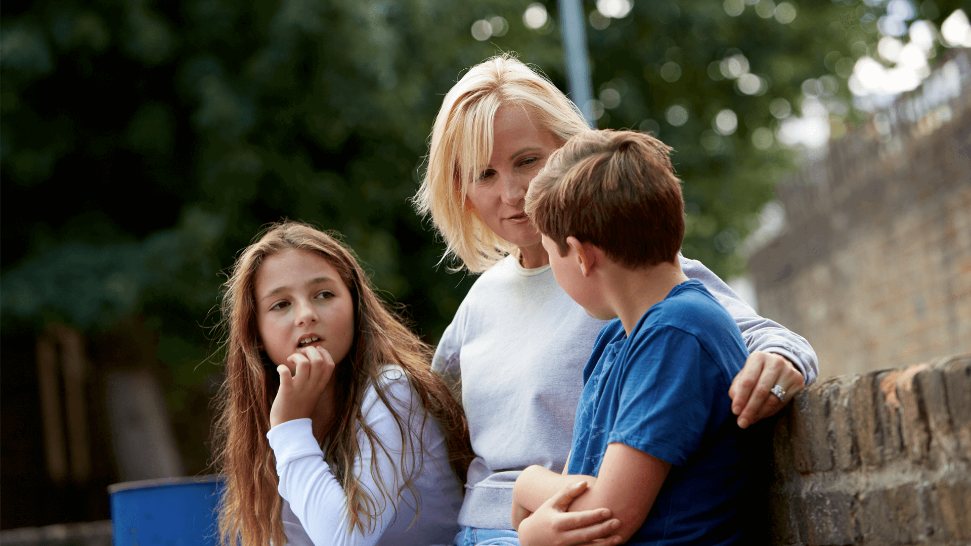 A mother sitting against a wall talking to her children with her arms around them