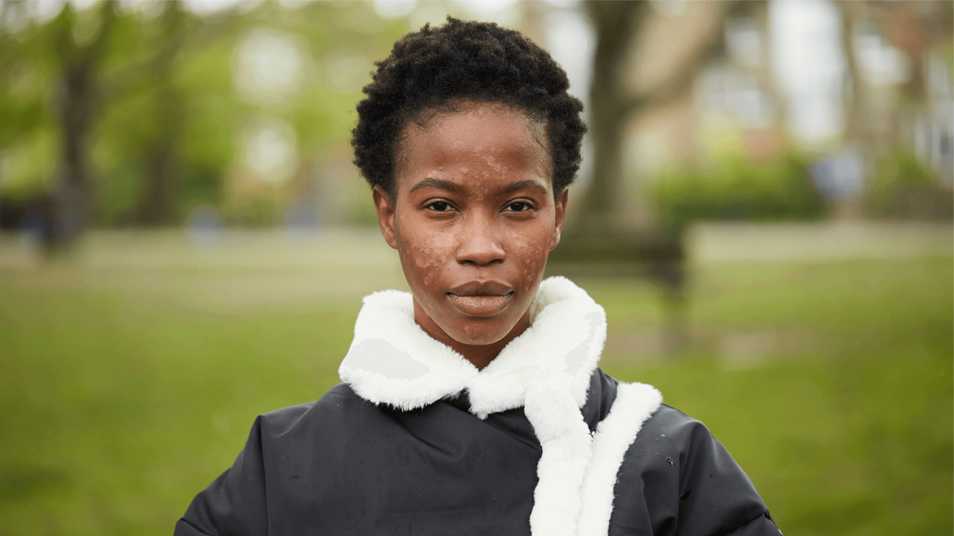 close-up-of-a-young-woman-with-short-curly-black-hair-and-in-black-jacket-looking-in-front-of-the-camera-with-trees-in-the-park-as-background