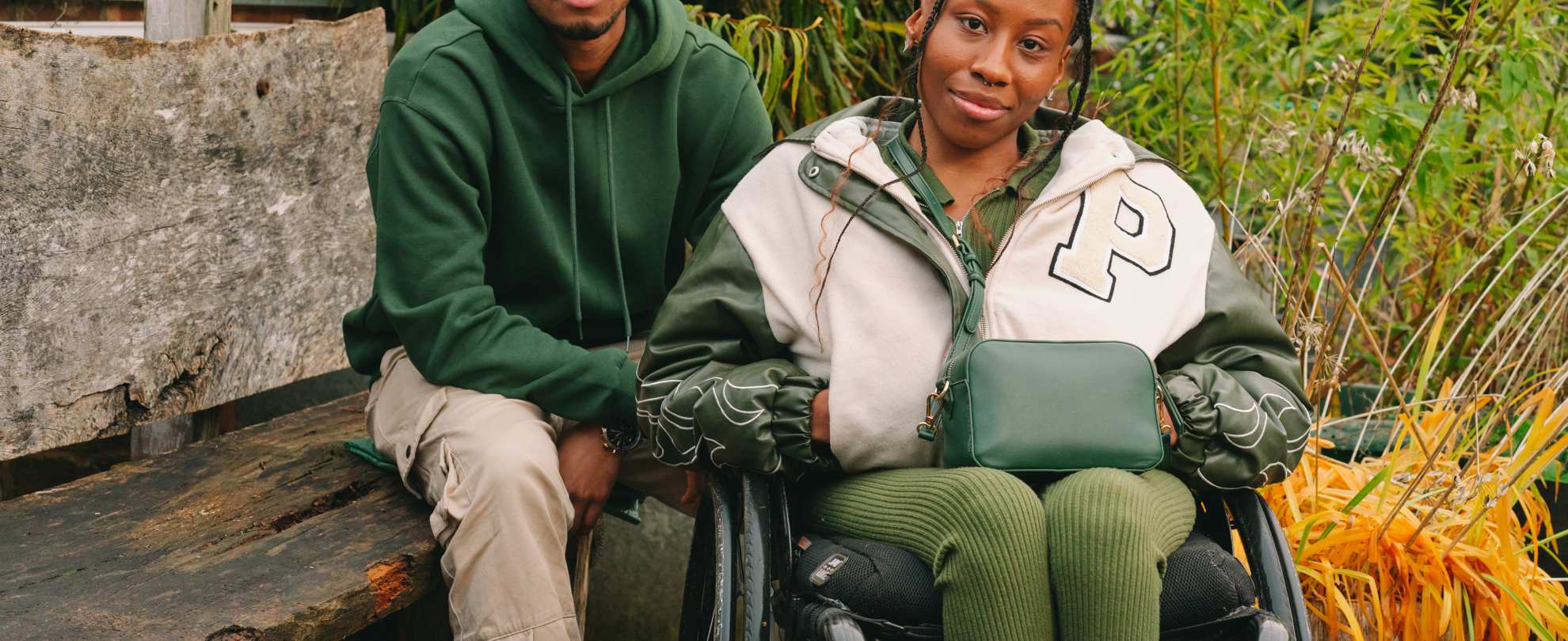 A young Black woman in a wheelchair and a young Black man on a bench, both staring at the camera looking serious.