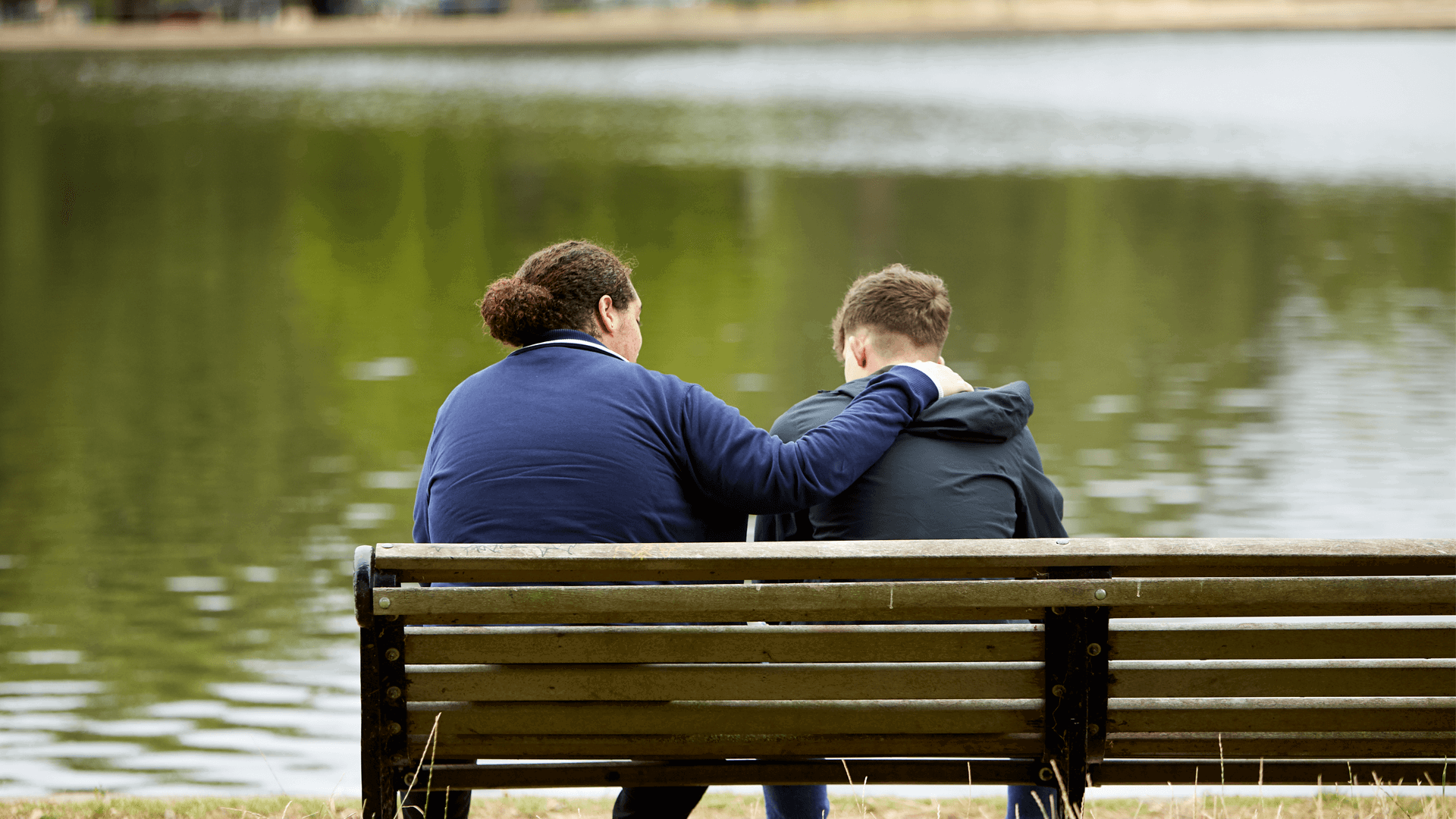 Two young people sitting on a bench in a park, one has their arm around the other.