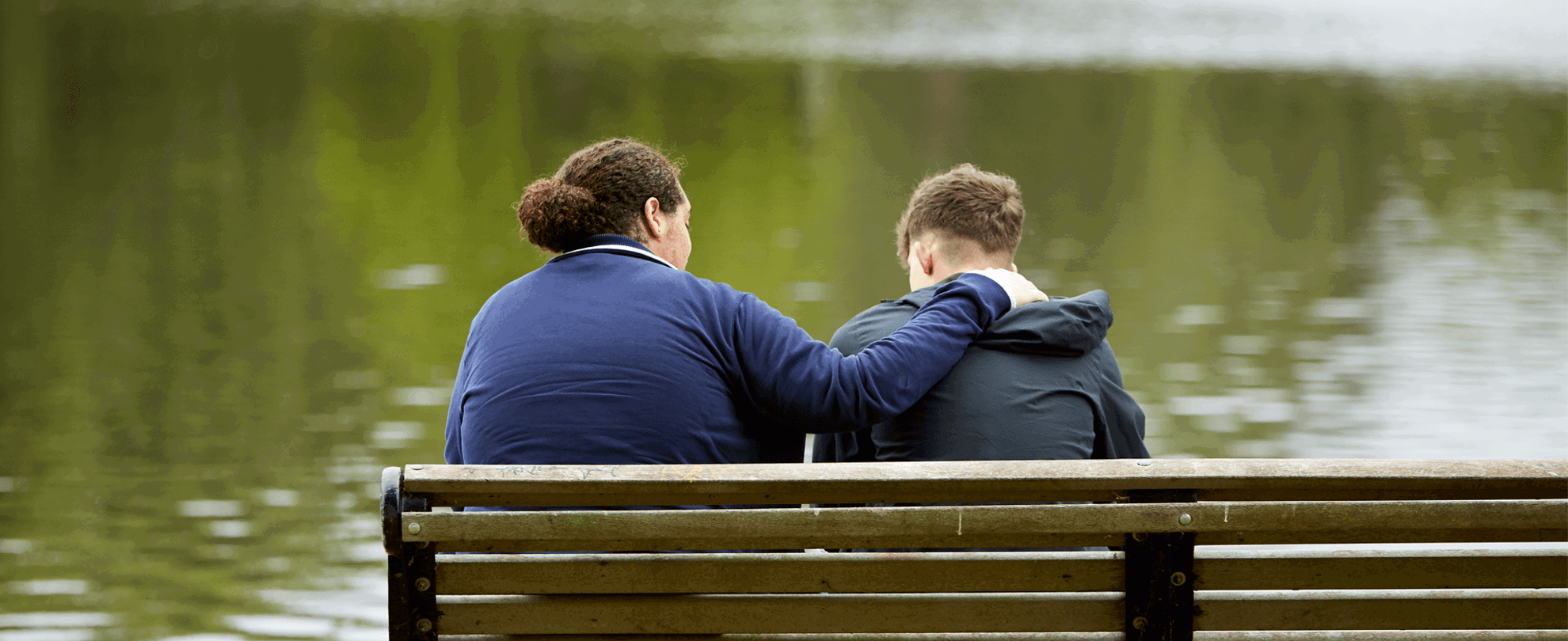 Two young people sitting on a bench in a park, one has their arm around the other.