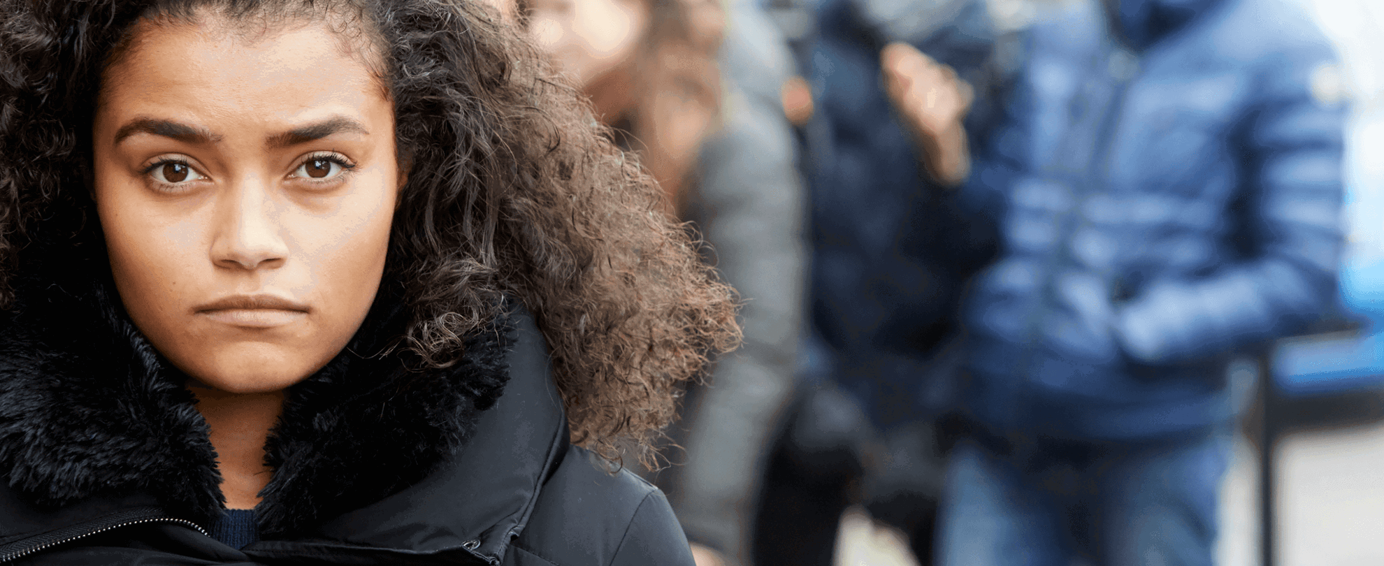 close up of a girl with a curly hair and wearing black jacket looking in front of the camera with group of young people on the background