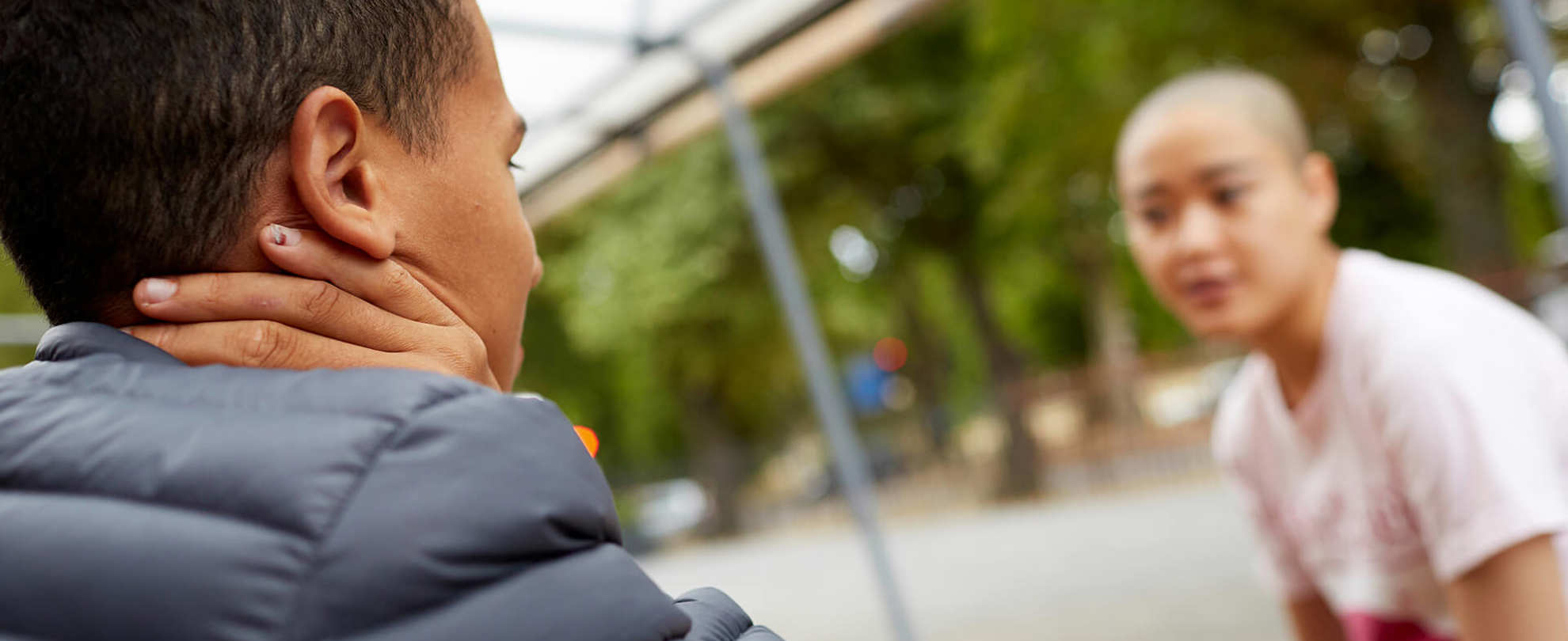 a-young-man-in-blue-jacket-with-face-unseen-talking-to-a-young-woman-with-shaved-head-in-white-shirt-while-sitting-on-a-bench-with-table-in-the-park