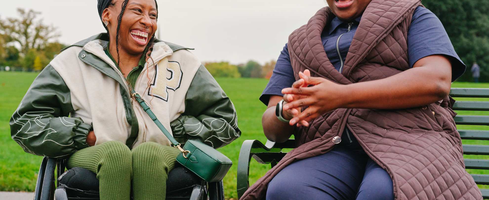 A young Black woman in a wheelchair and an older Black woman sitting on a bench in the park. They are laughing together.