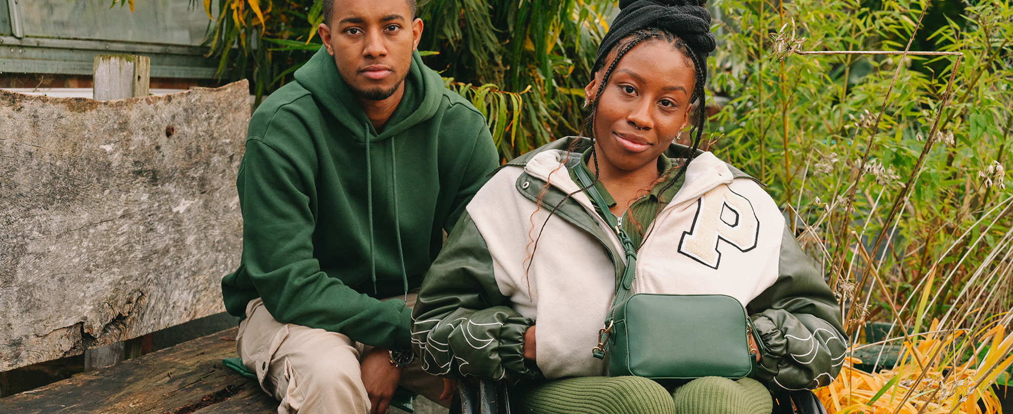 A young Black woman in a wheelchair and a young Black man on a bench, both staring at the camera looking serious.