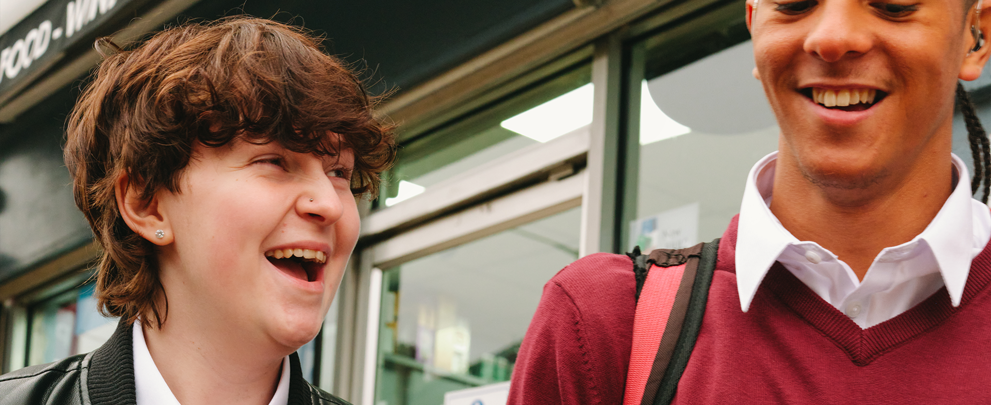 A Black teenage boy wearing a hearing aid laughing with a white non-binary teenager outside the shops.