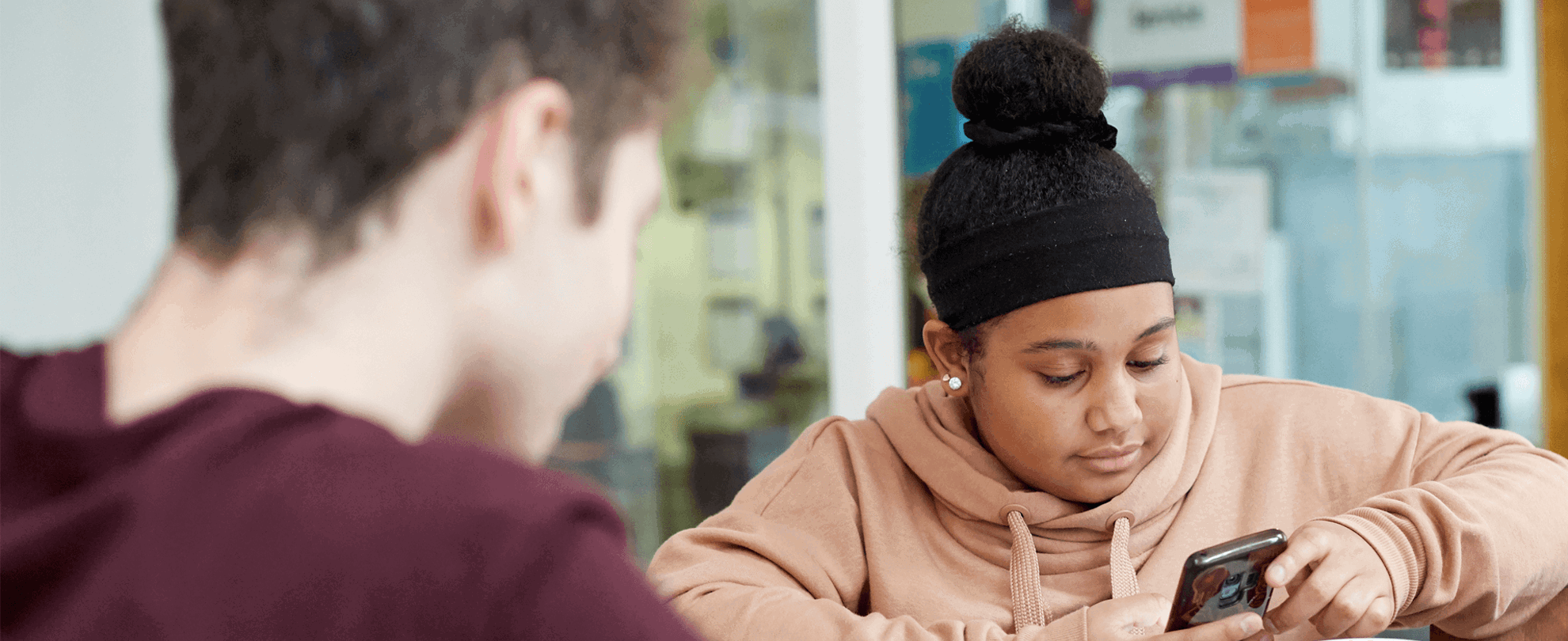 a girl wearing pink hoodie and a boy wearing red shirt in front of her are busy looking at their phones while sitting inside the campus