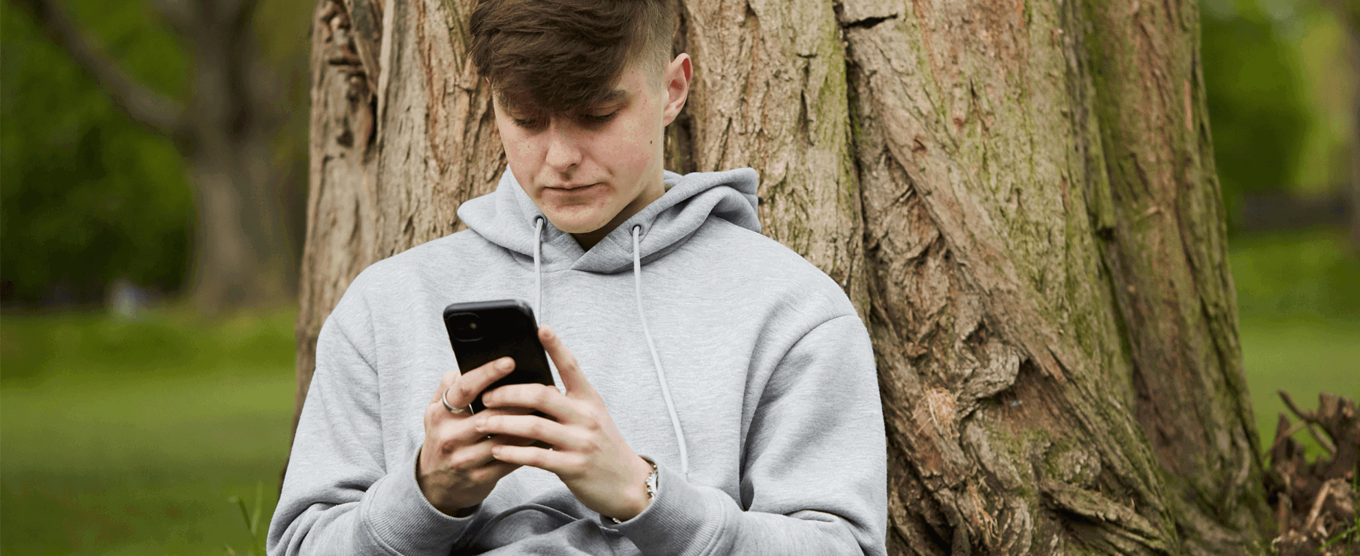 a boy wearing grey hoodie using his mobile phone while sitting on the ground leaning on a tree