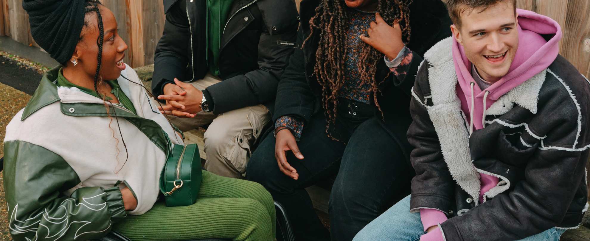 A group of young people laughing together outside on a bench. Group includes two Black young women (one in a wheelchair), one Black young man, and a white young man.