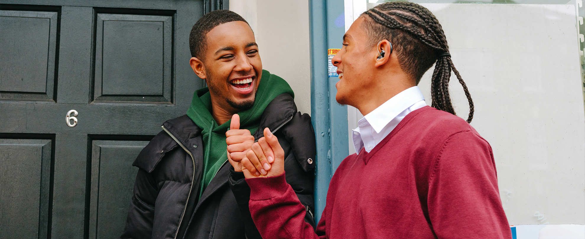 A Black teenage boy wearing a hearing aid bumping fists with a young Black man outside a front door.