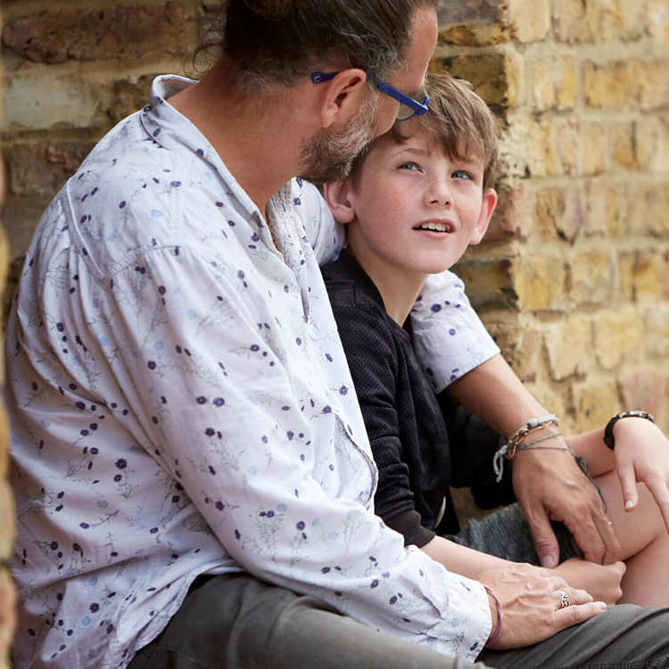 A father with his arm around his son sitting by a wall.