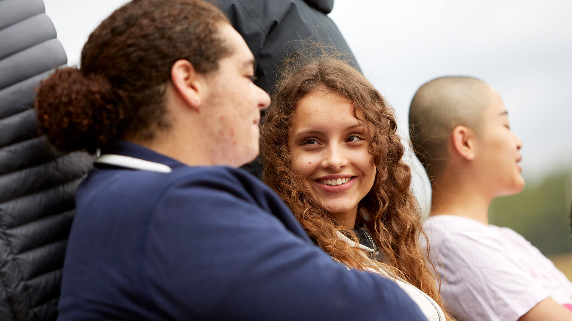Three young people sit on a bench outside. The person on the right is chatting with the person in the middle of the bench who is looking at them and smiling. The person on the right is speaking to someone outside of the image.