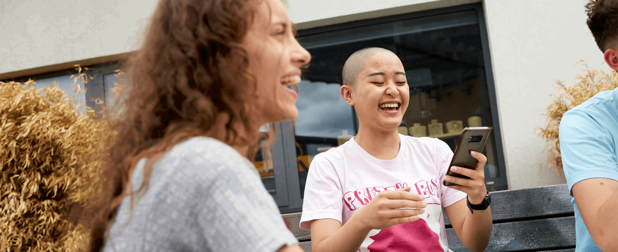 Two girls sitting together at a park bench with their friends. They are both laughing and one is looking at her phone.