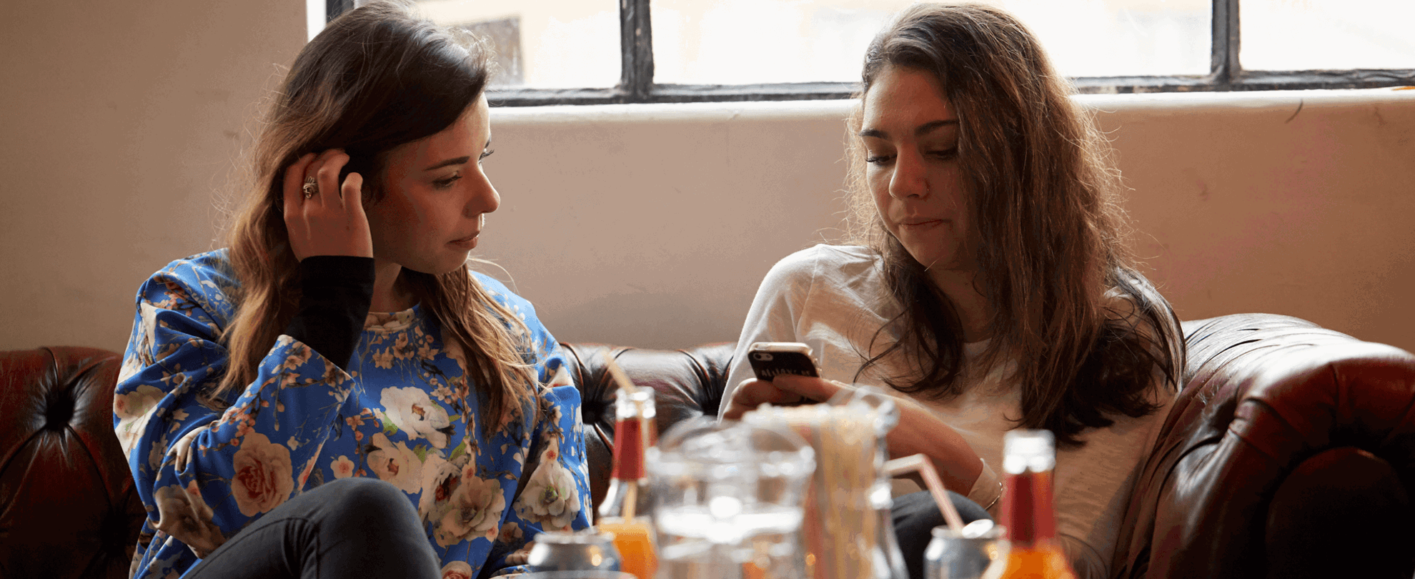 medium shot of two girls sitting on a couch one is using a phone while another girl is glancing on her phone bottles and drinks are on the table as foreground