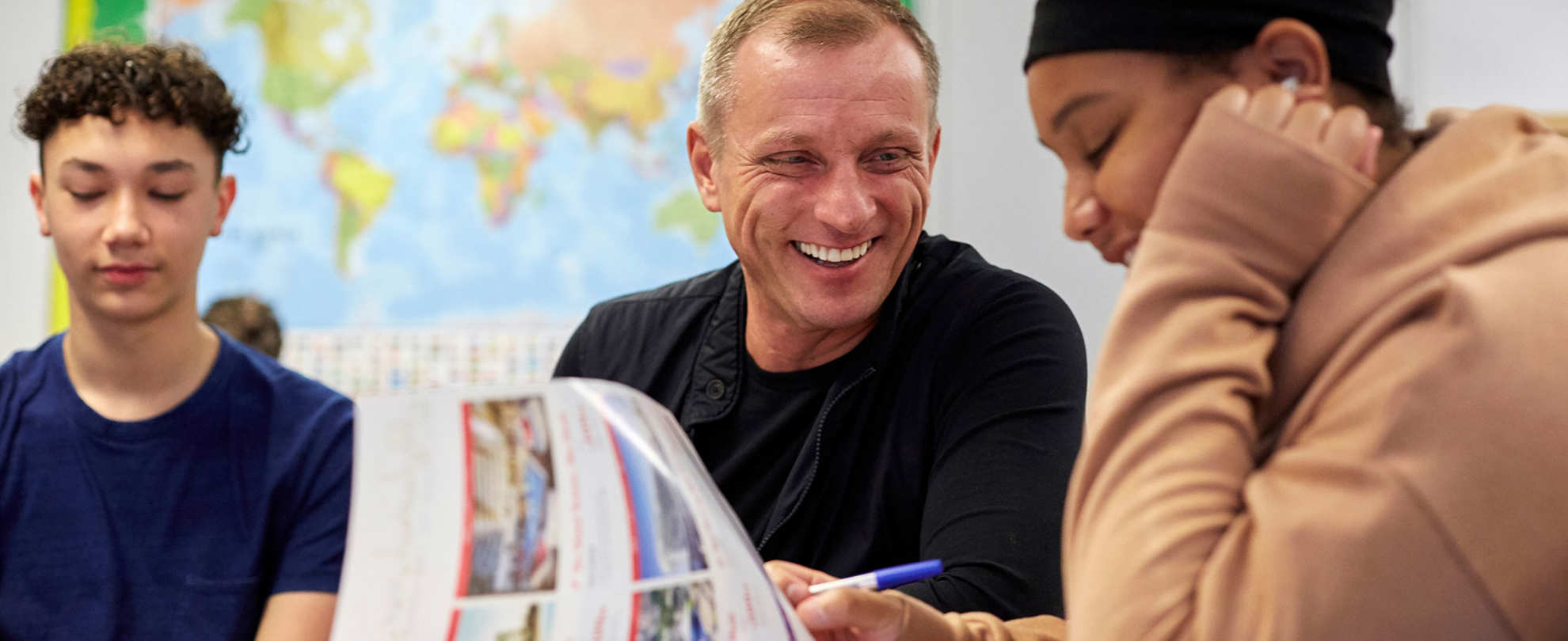 a teacher is laughing while he sits in between two of his students who are also laughing inside the classroom