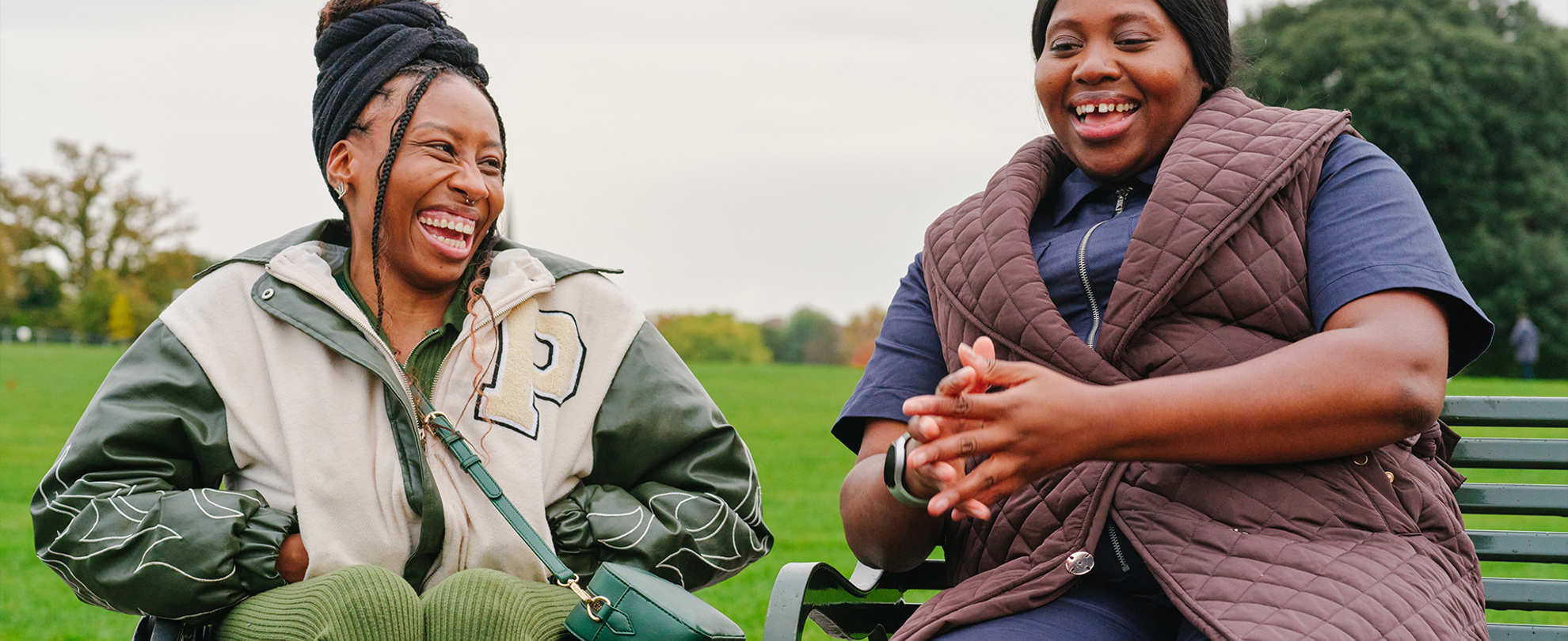 A young Black woman in a wheelchair and an older Black woman sitting on a bench in the park. They are laughing together.