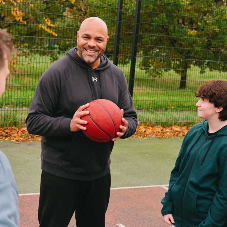 A group of young people playing basketball with an older Black man. The group of young people includes: one white young man, one white non-binary teenager and one Black young woman.