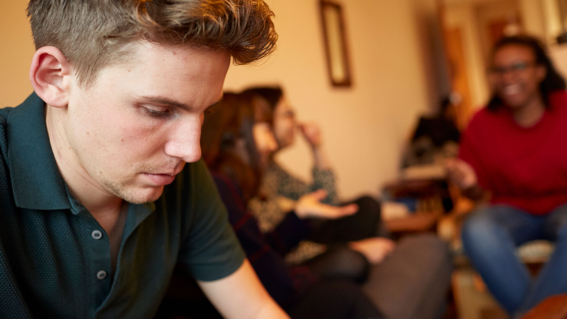 A young man looks worried and alone, while his three friends are talking and laughing in the background.