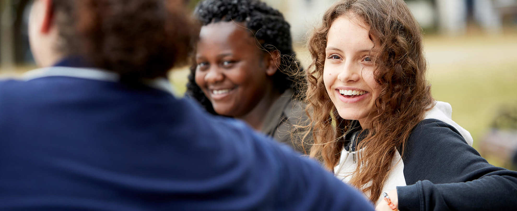 Three young people sit on a bench talking and laughing. The person on the left has their back to the camera and wears a dark blue jacket. The person in the middle has long wavy hair and wears a black and white jacket. The person on the right has curly hair and wears a green jacket.