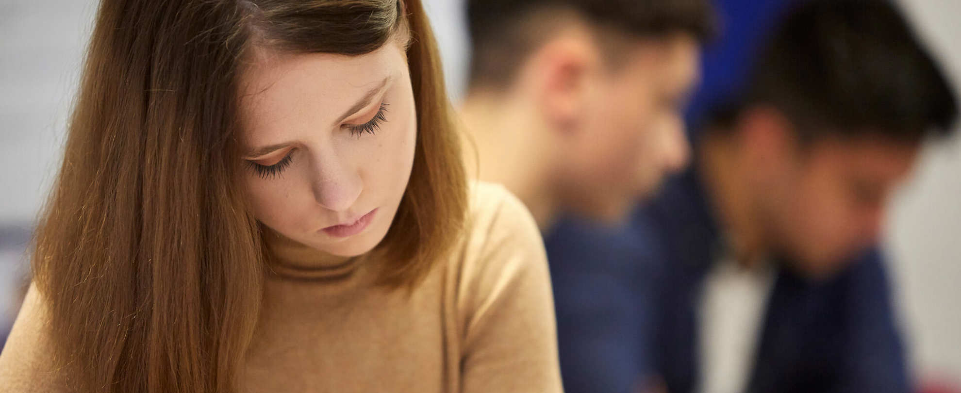 A young woman in a brown jumper looking down with a sad expression with two young men in the background.