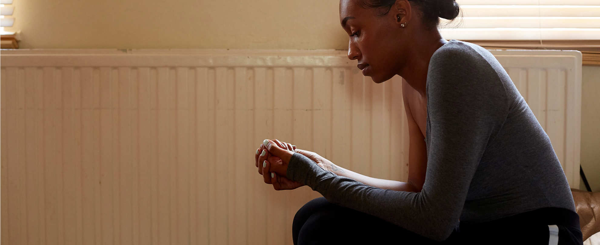 A girl sitting on a sofa in a living room beside a radiator. She is hunched over and looking down at her hands clasped together.