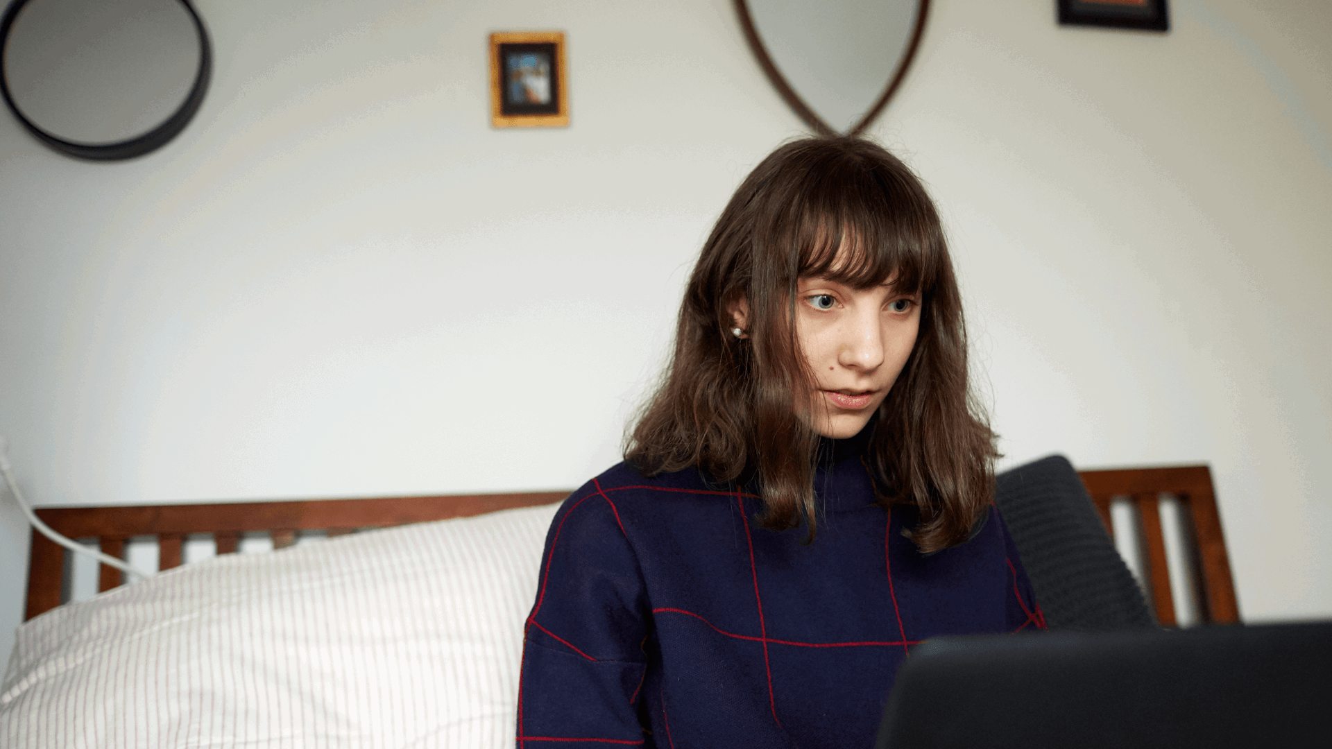 A girl sits on the end of her bed while looking at her laptop.