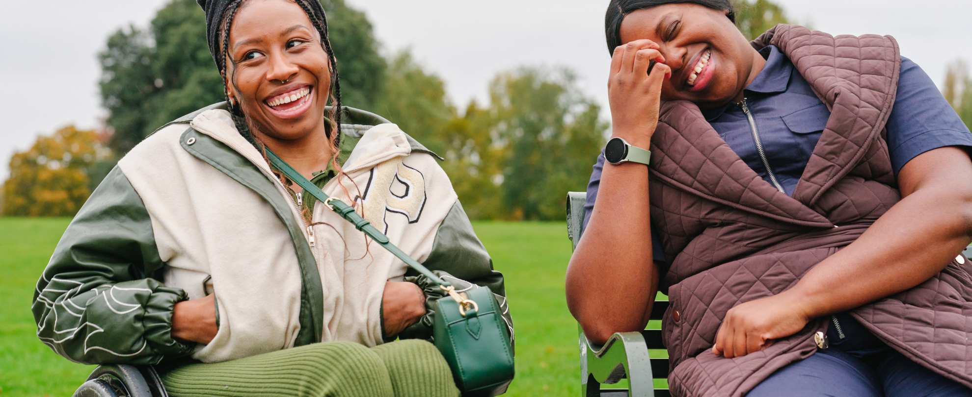 A young Black woman in a wheelchair and an older Black woman sitting on a bench in the park. They are laughing together.