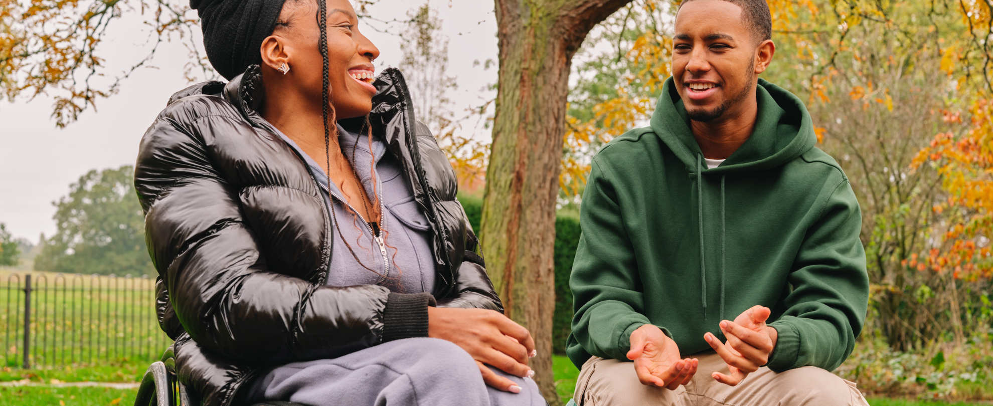 A young Black woman in a wheelchair talking to a young Black man on a bench in the park. The woman is laughing while the man explains something.