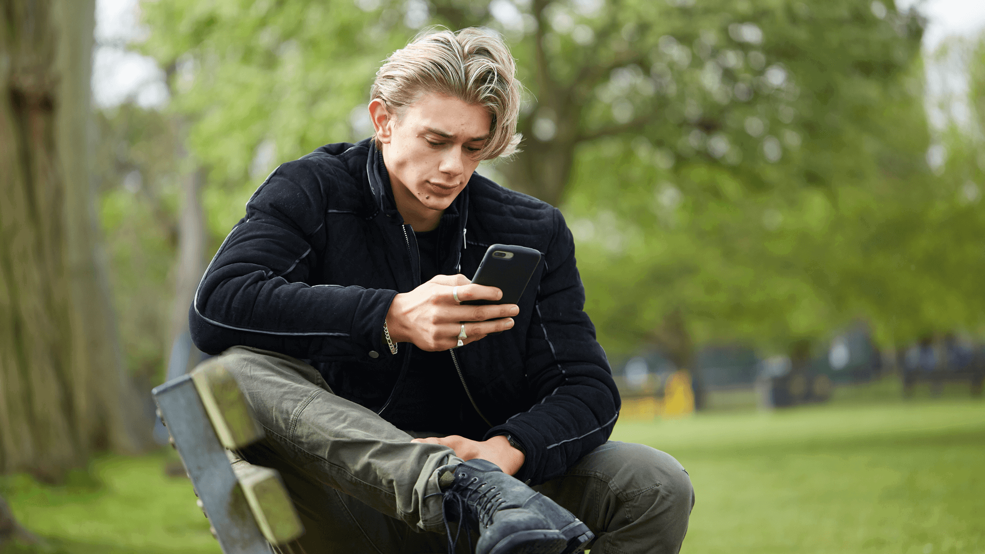 A young man wearing a black jacket sits on a park bench. He is looking at his phone with a worried expression.