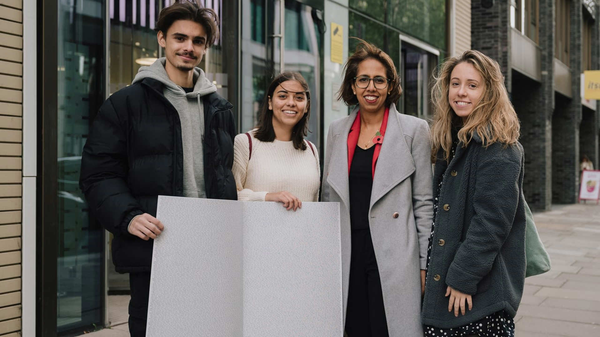 Three YoungMinds Activists with MP Munira Wilson holding the End The Wait card open on a page with all the signatures.