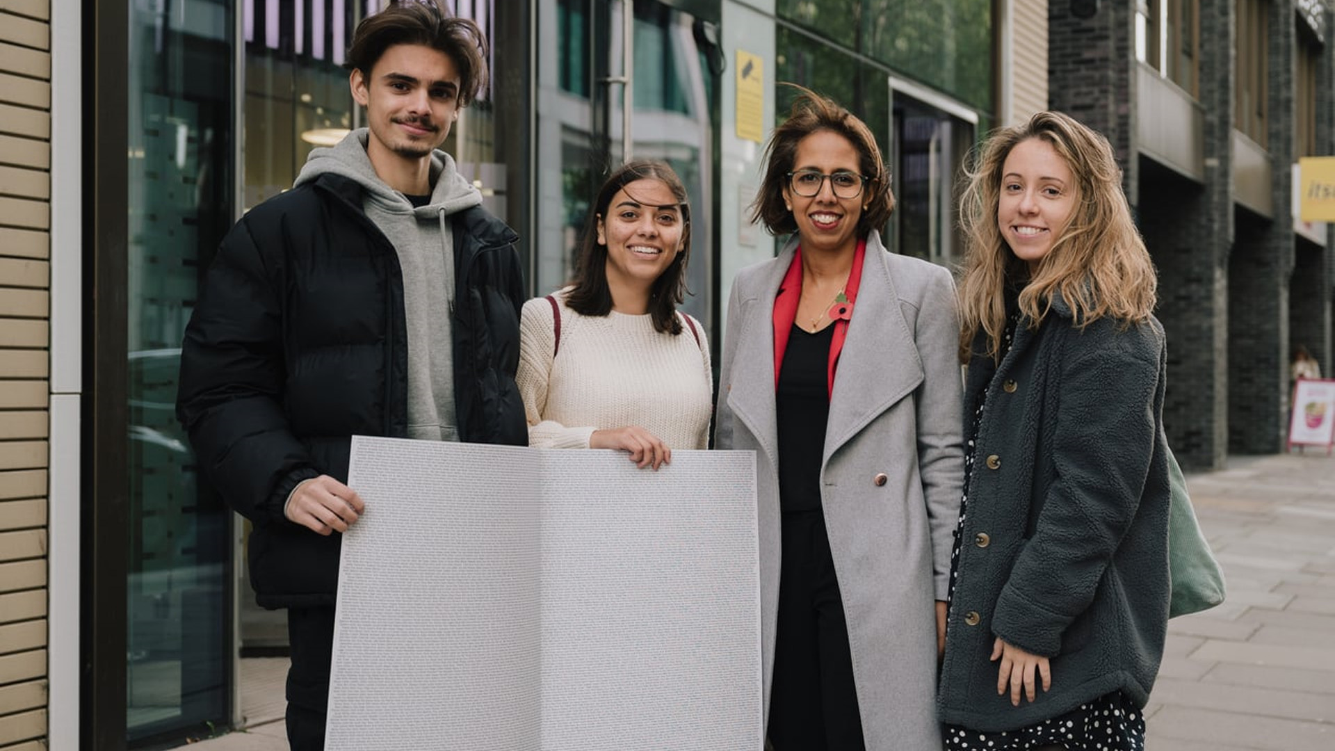 Three YoungMinds Activists with MP Munira Wilson holding the End The Wait card open on a page with all the signatures.