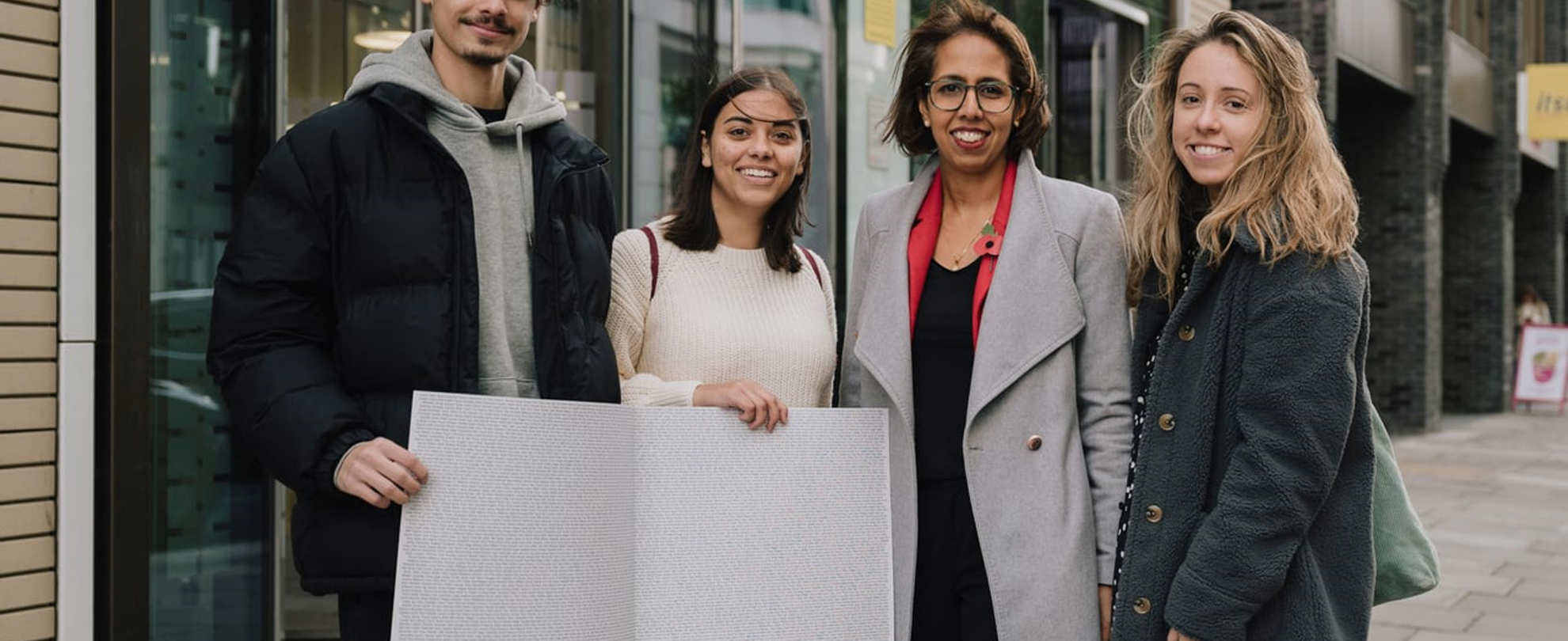 Three YoungMinds Activists with MP Munira Wilson holding the End The Wait card open on a page with all the signatures.