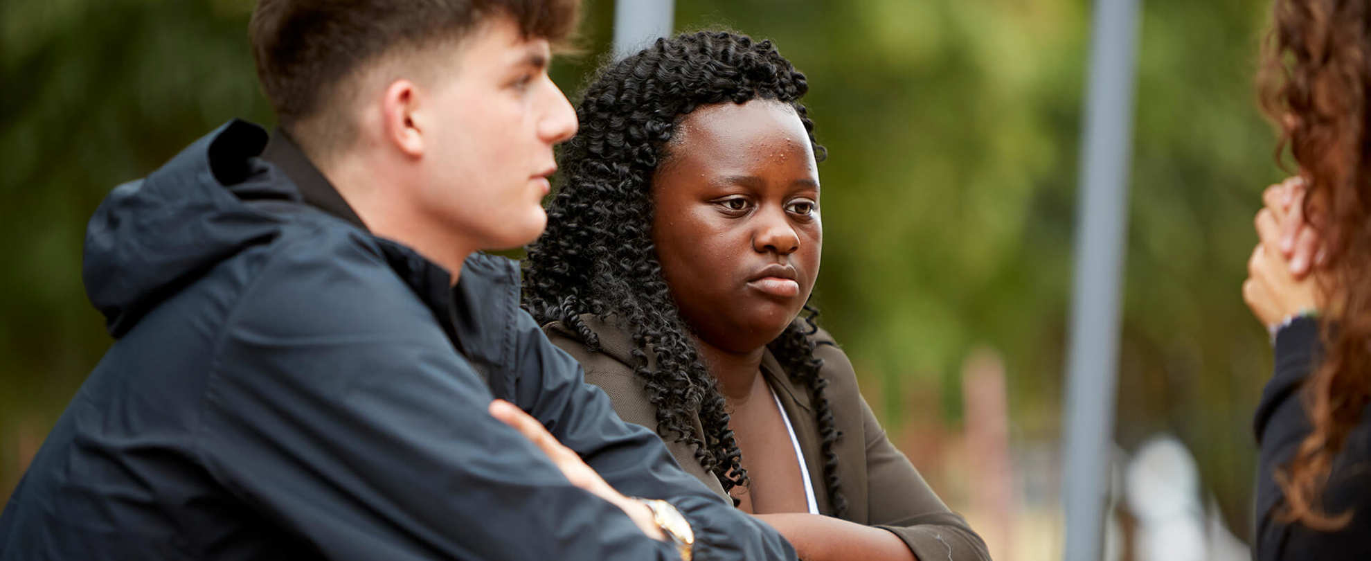 close-up-of-a-young-girl-with-curly-hair-and-a-boy-in-blue-jacket-looking-serious-at-another-girl-cropped-in-the-photo-while-sitting-on-a-bench-with-table-in-the-park