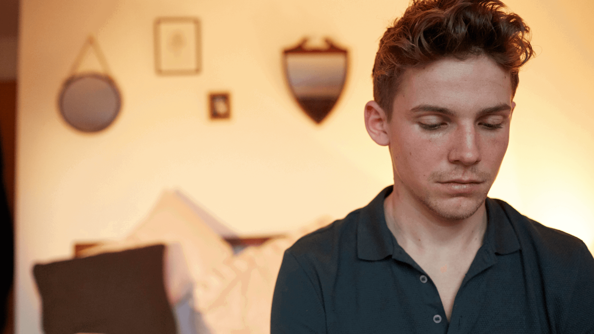 A boy sitting alone in his bedroom. He looks sad with his head and eyes facing down.