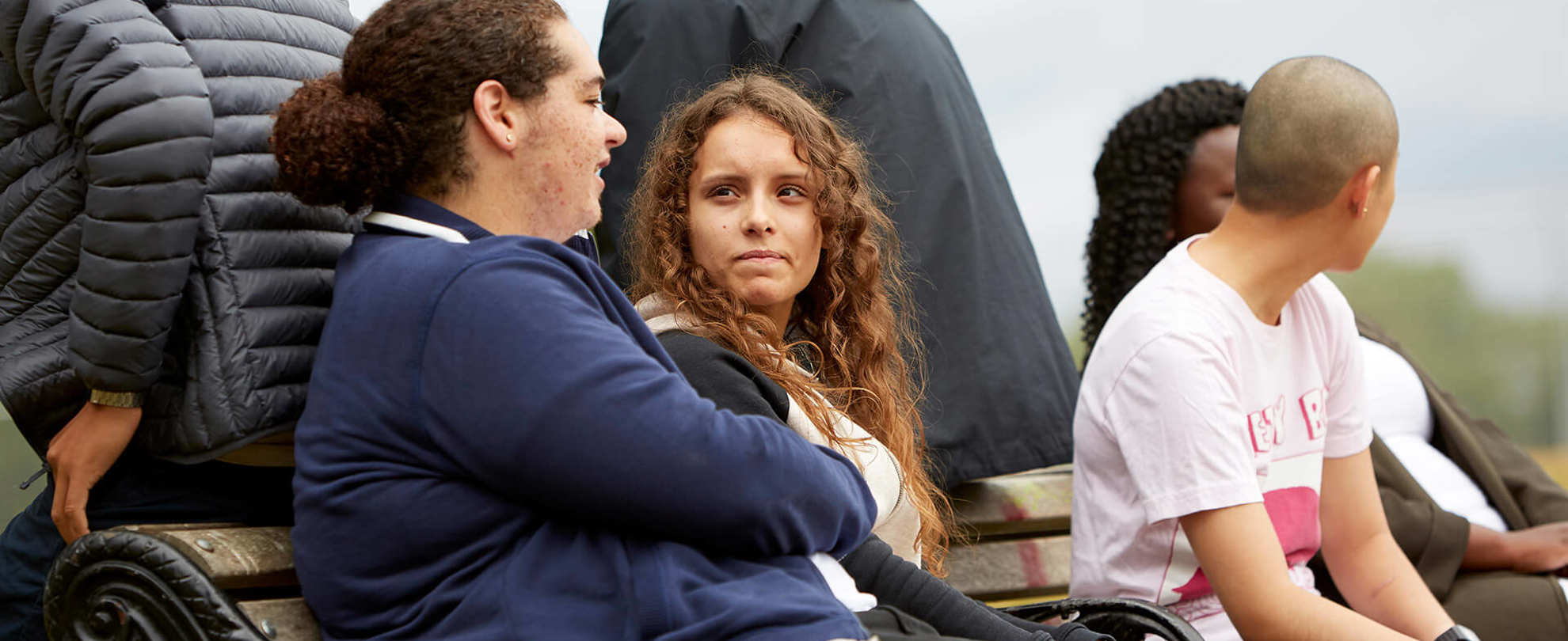 A group of six young people sitting on a bench and talking to each other in a park.