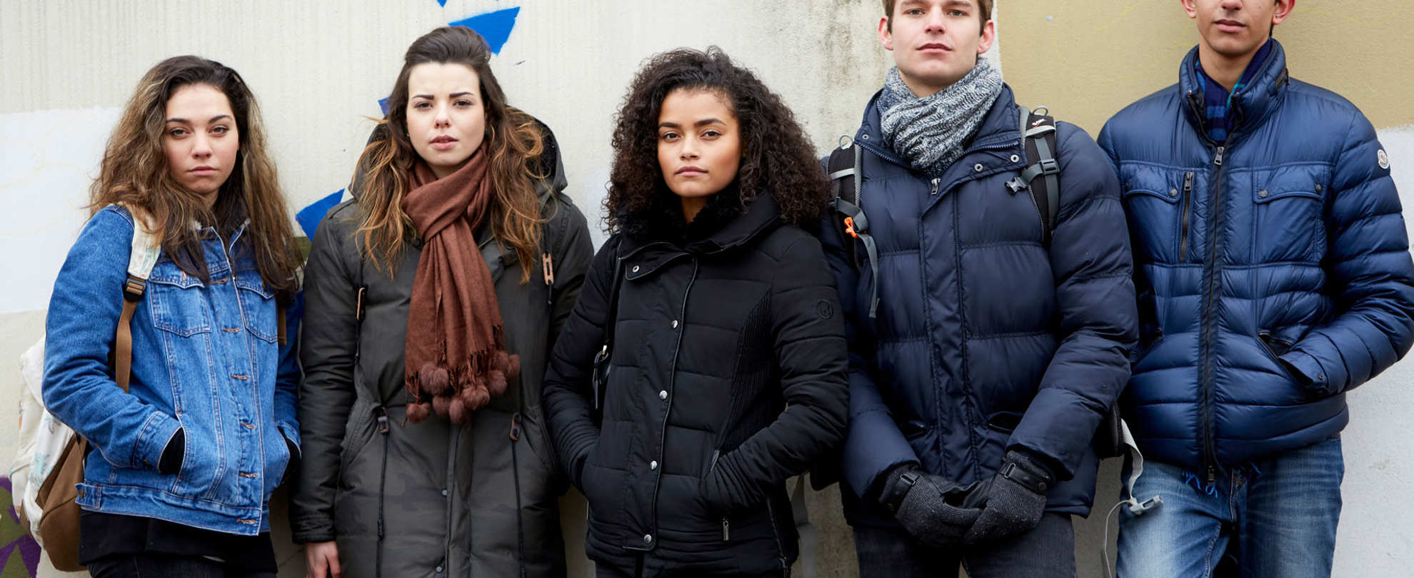 A group of five young people wearing jackets, standing side by side against a wall and looking straight ahead.